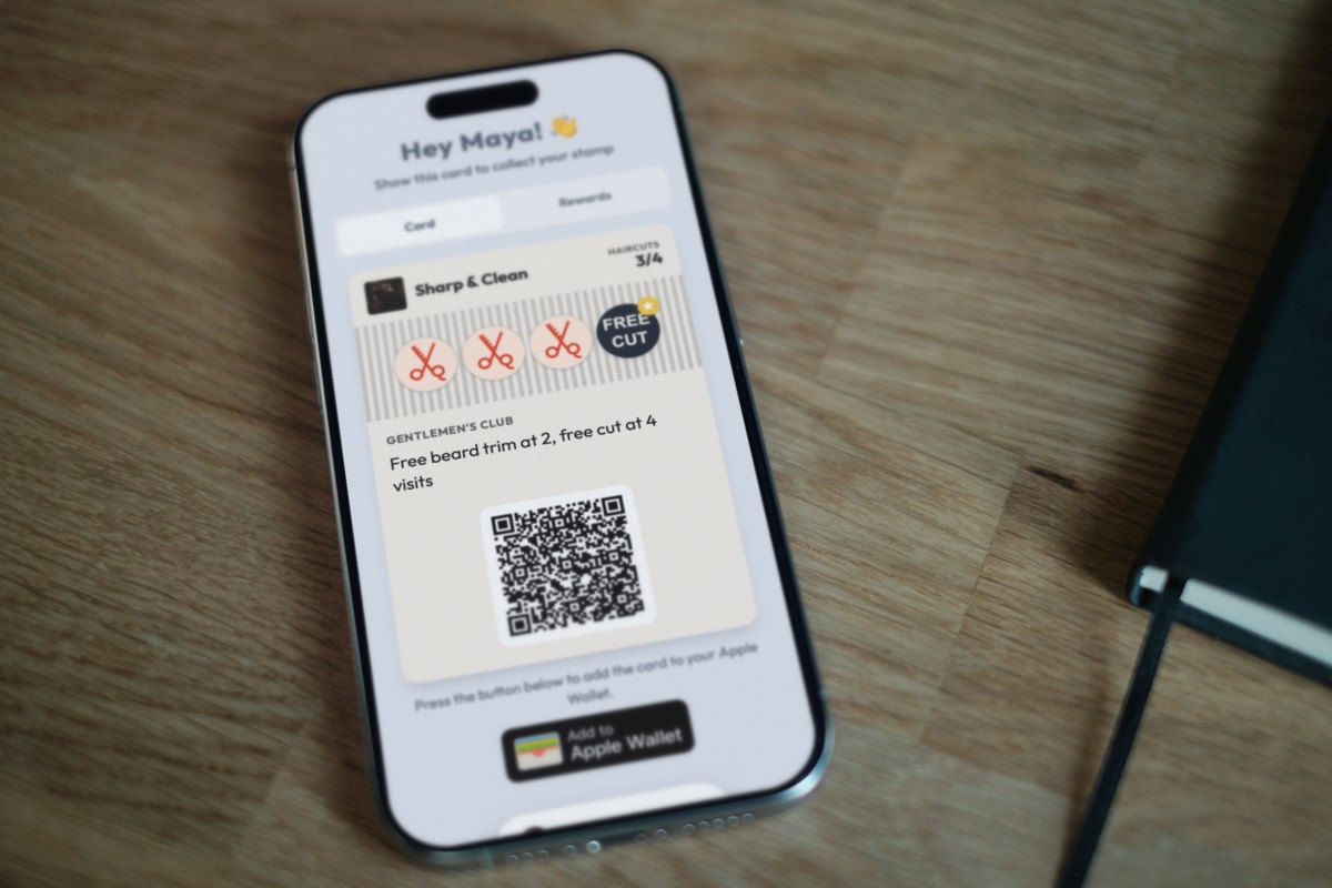 Illustration: an iPhone lying on a wooden table next to a black notebook, showing a Sharp & Clean barber loyalty card at 3 of 4 haircuts with the line 'Free beard trim at 2, free cut at 4 visits' and an Add to Apple Wallet button