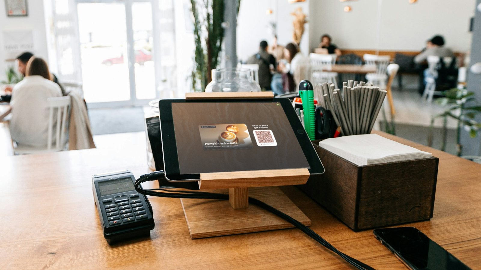 Illustration: a café counter with a card terminal next to an iPad on a wooden stand showing a loyalty sign-up screen with a QR code