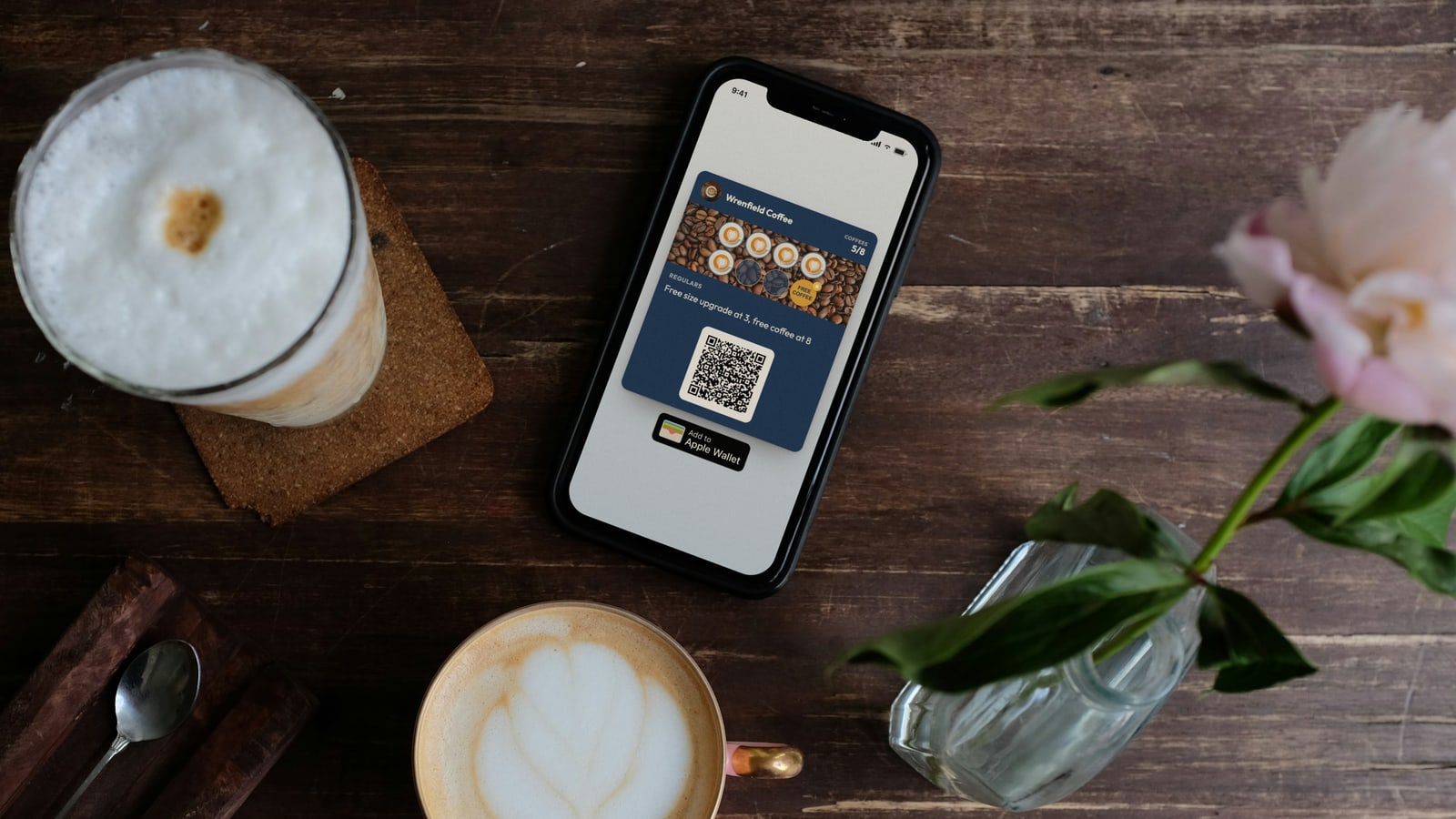 Illustration: an iPhone on a café table showing a Wrenfield Coffee loyalty card at 5 of 8 coffees, with a QR code and an Add to Apple Wallet button