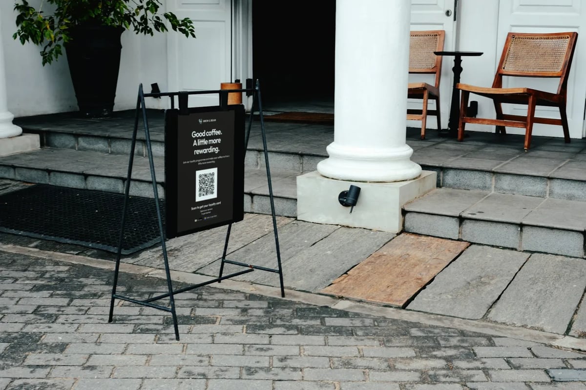 Illustration: a black A-frame sign on the pavement outside a café entrance holds a printed poster with a QR code and the words Scan to get your loyalty card