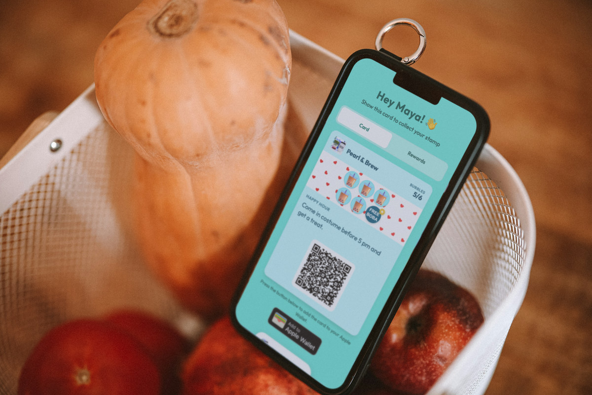 Illustration: a phone leaning in a wire basket with a butternut squash and red apples, showing a Pearl & Brew bubble tea loyalty card during a Happy Hour campaign with the line 'Come in costume before 5 pm and get a treat.'