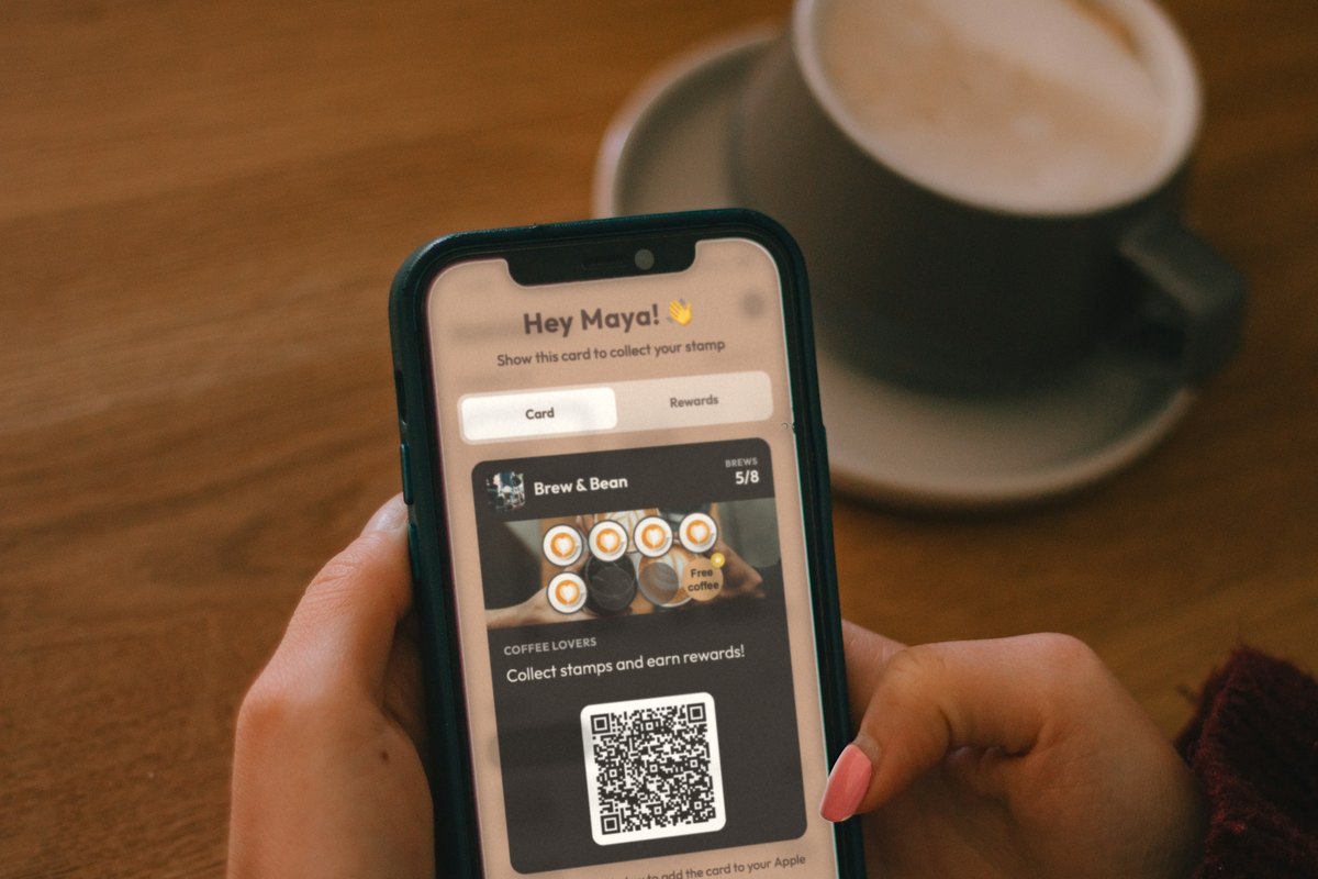 Illustration: hands holding a phone over a café table next to a cup of coffee, showing a Brew & Bean loyalty card with 5 of 8 coffee stamps collected and a free coffee on the last stamp