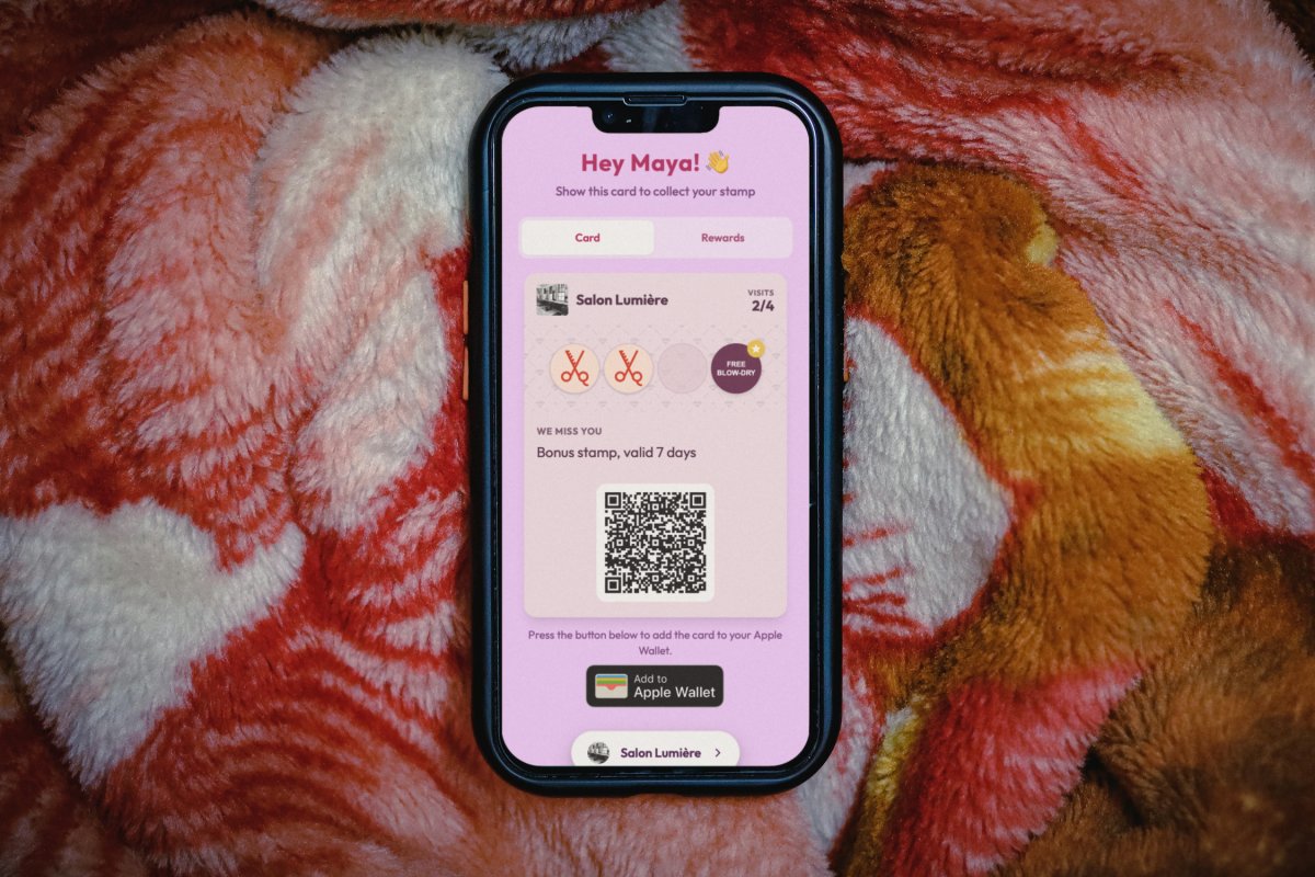 Illustration: an iPhone lying on a red and white fleece blanket, showing a Salon Lumière loyalty card page at 2 of 4 visits with the campaign header WE MISS YOU and the line Bonus stamp, valid 7 days