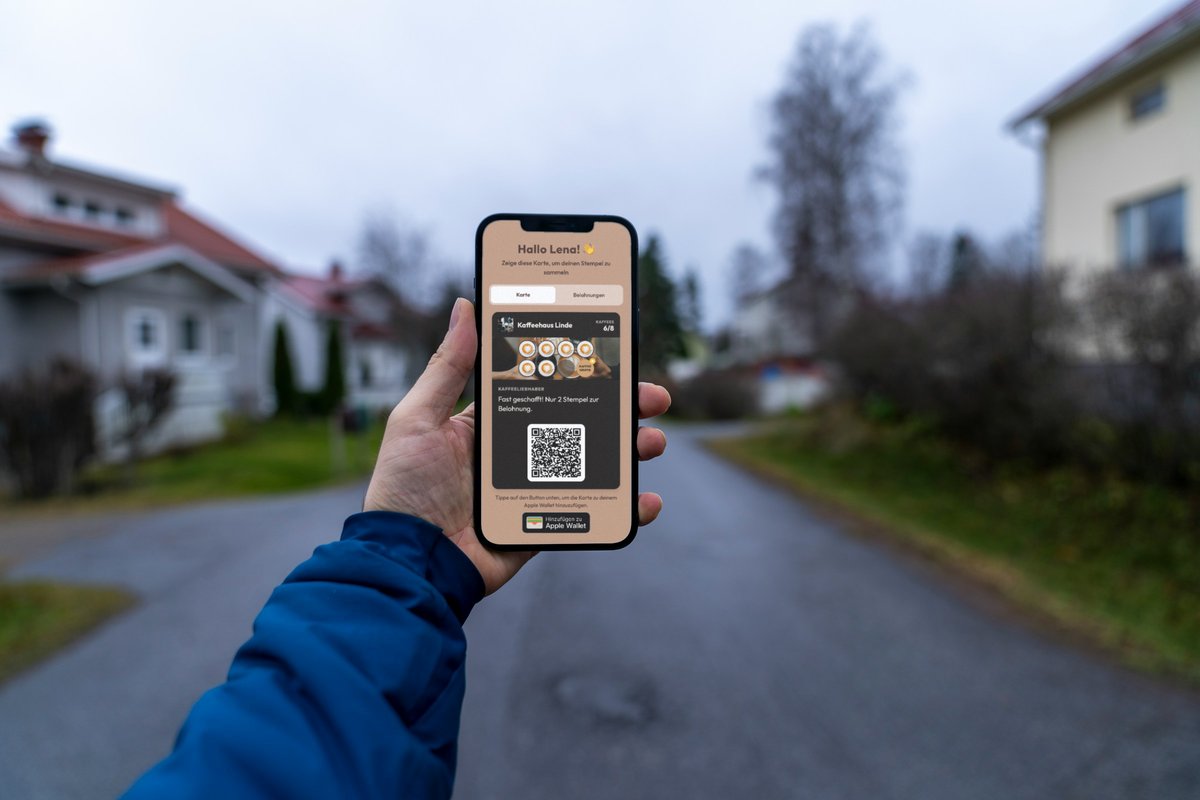 Illustration: eine Hand im blauen Jackenärmel hält in einer ruhigen Wohnstraße ein Smartphone hoch; auf dem Display die Treuekarte vom Kaffeehaus Linde mit 6 von 8 Kaffees mit dem Hinweis „Fast geschafft! Nur 2 Stempel zur Belohnung.“