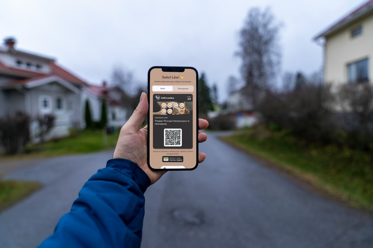 Illustration : une main dans une manche de doudoune bleue tient un téléphone dans une rue résidentielle calme ; l'écran affiche la carte de fidélité de Café Lumière à 6 cafés sur 8 avec la phrase « Presque ! Plus que 2 tampons pour la récompense. »