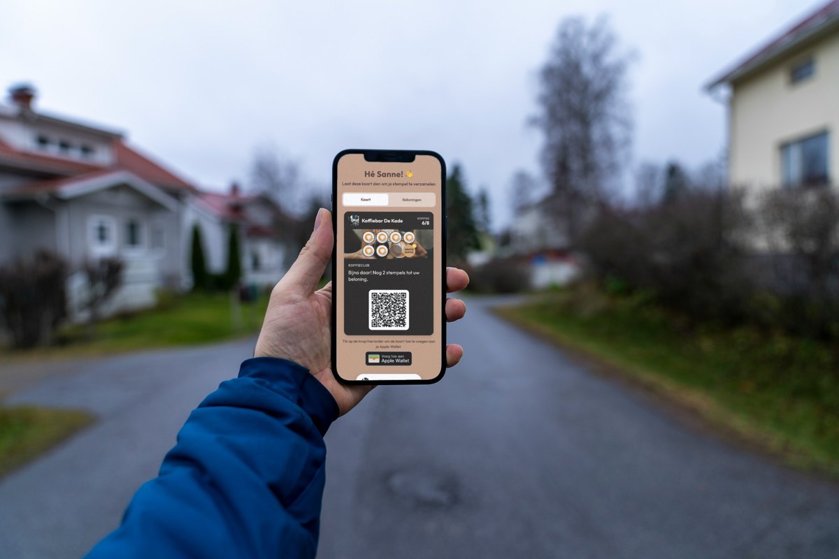 Illustratie: een hand in een blauwe jas houdt een telefoon omhoog in een rustige woonstraat; op het scherm de klantenkaart van Koffiebar De Kade op 6 van de 8 koffies met de regel "Bijna daar! Nog 2 stempels tot uw beloning."