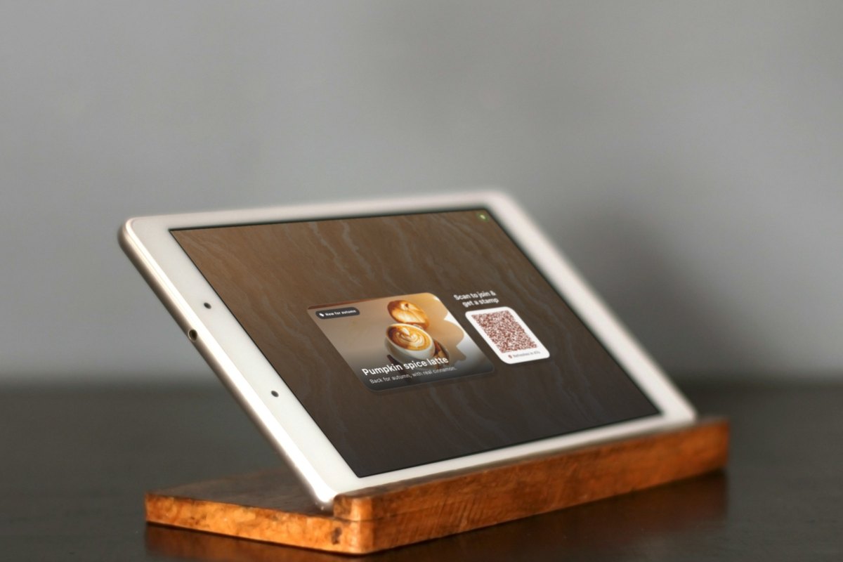 Illustration: close-up of an iPad on a wooden stand showing a pumpkin spice latte promotion and a QR code to join and get a stamp