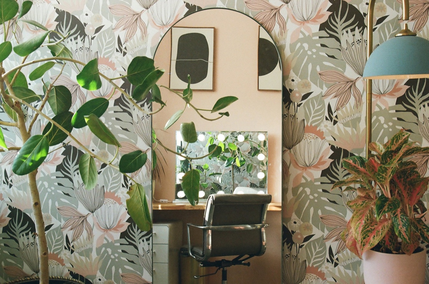 A lash studio corner: an arched mirror reflecting a chair and a lit vanity mirror, with botanical wallpaper and plants
