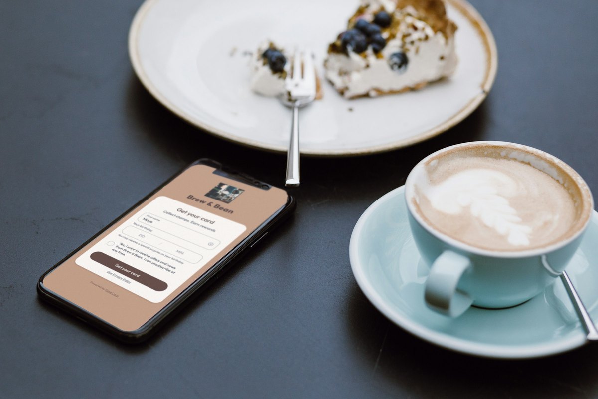 Illustration: an iPhone lying on a dark café table next to a latte and a slice of blueberry cheesecake, showing a café's loyalty card sign-up form with the first name Maya typed in and a Get your card button