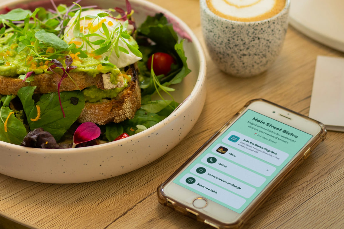 Illustration: a phone lies on a café table beside an avocado-toast bowl and a latte, showing the Main Street Bistro business page: "Neighborhood bistro for weeknight dinners and weekend brunch.", 214 Main Street, a "Join the Bistro Regulars" loyalty card button (free dessert on the 2nd visit, a free entrée on the 6th), then Menu, Call us, Leave a review on Google and Reserve a table.