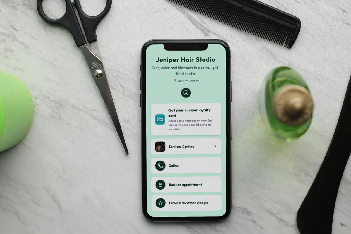 Illustration: a phone lies on a marble salon counter between hair-cutting scissors and a comb, showing the Juniper Hair Studio business page: "Cuts, color and blowouts in a calm, light-filled studio.", 48 Elm Street, an Instagram button, a "Get your Juniper loyalty card" button (a free scalp massage on the 2nd visit, a free deep conditioning on the 4th), then Services & prices, Call us, Book an appointment and Leave a review on Google.