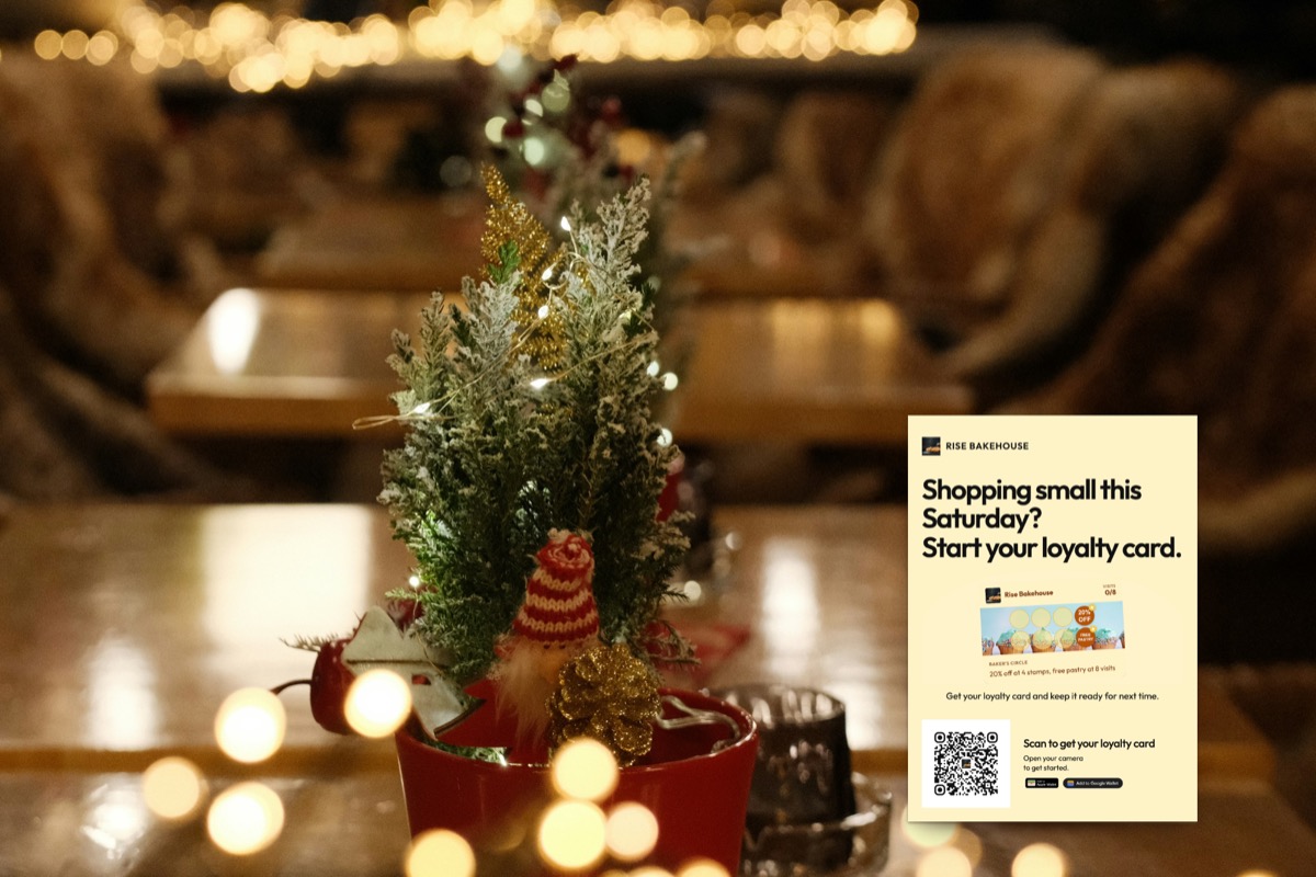 Illustration: an A6 loyalty card sign reading 'Shopping small this Saturday? Start your loyalty card.' on a café table next to a small decorated Christmas tree