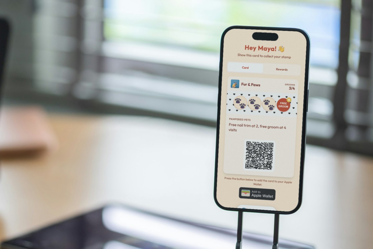 Illustration: a phone in a small stand on a desk by a window, showing a Fur & Paws pet groomer loyalty card at 3 of 4 grooms with the line 'Free nail trim at 2, free groom at 4 visits' and a QR code