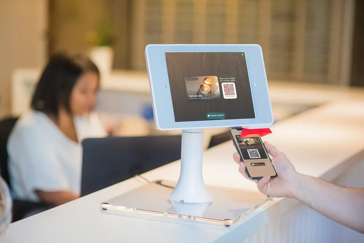 Ilustrace: ruka drží telefon s věrnostní kartou a konfetami vedle kiosku na iPadu s nápisem "Stamp collected!".