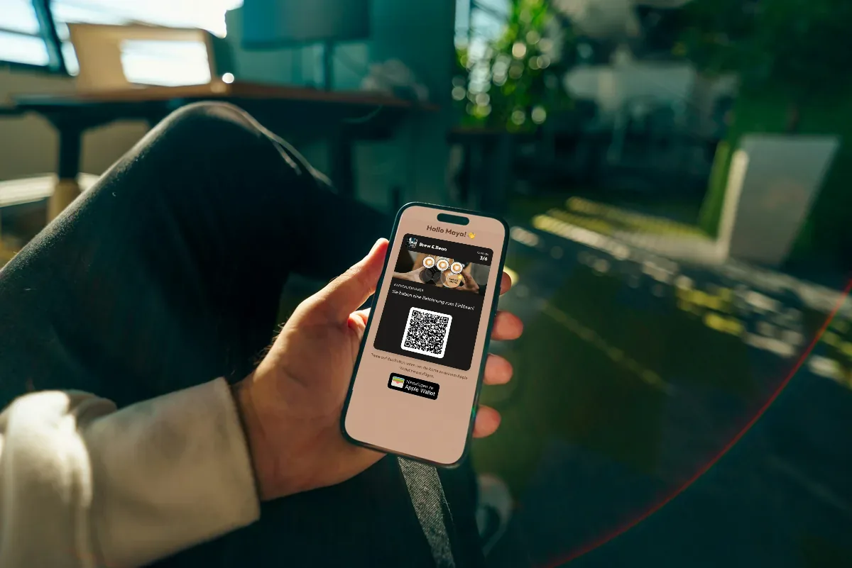 Illustration: eine Hand hält ein Handy mit der Treuekarte eines Cafés, Sternkonfetti und einer Schaltfläche zum Hinzufügen zu Apple Wallet.