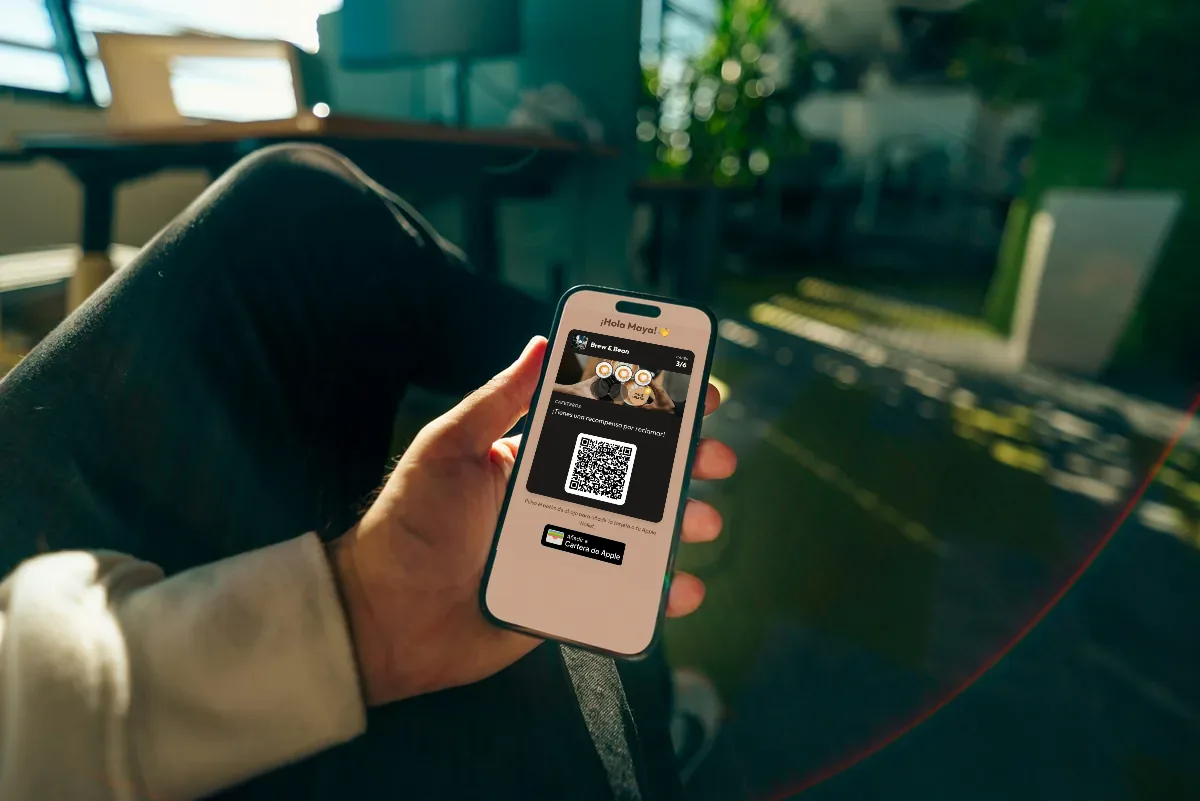 Ilustración: una mano sostiene un móvil con la tarjeta de fidelidad de una cafetería, confeti de estrellas y un botón para añadirla a Apple Wallet.